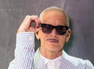 photograph of john waters, an older white man with a small moustache, wearing sunglasses and a seersucker suit