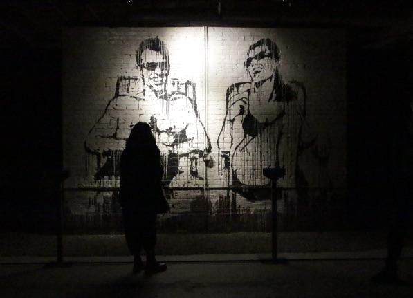 Installation view of Banksy: Genius or Vandal? featuring Stop Esso. The artwork is painted onto a brick wall that has been removed from the original building and installed as an isolated slab of brick in a gallery