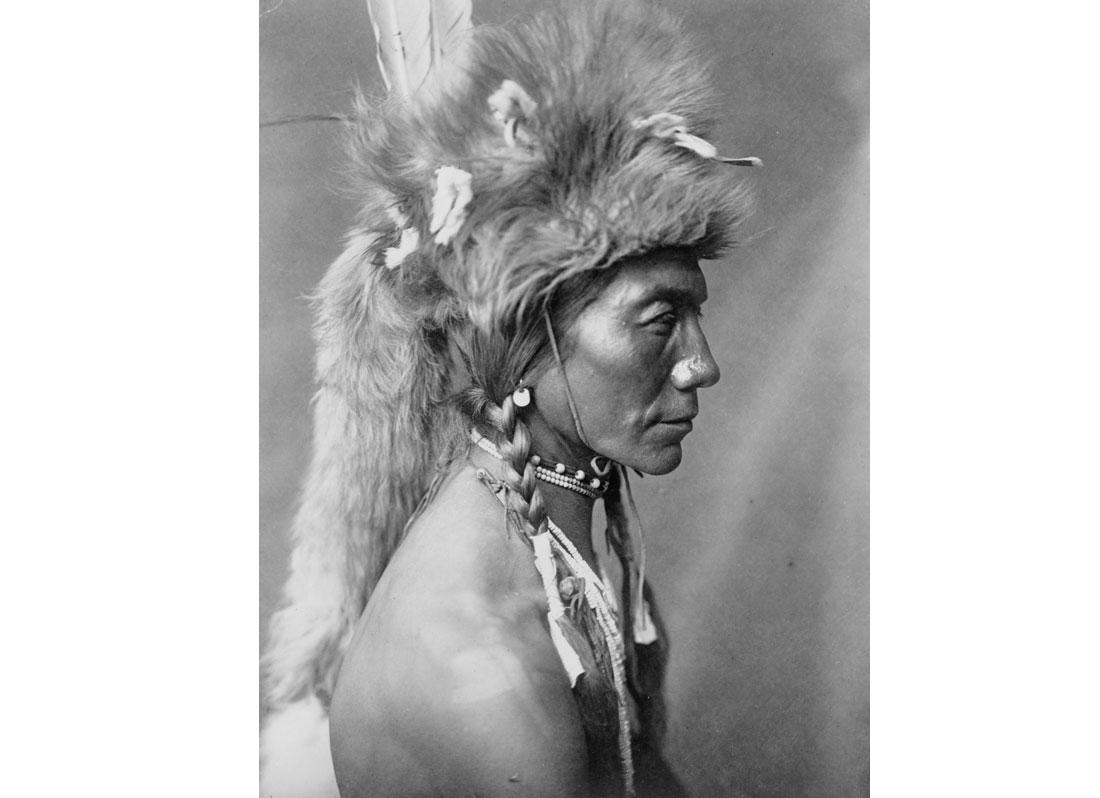 Yellow Kidney, Piegan man, head-and-shoulders portrait, facing left, wearing a fur headdress, 1910.