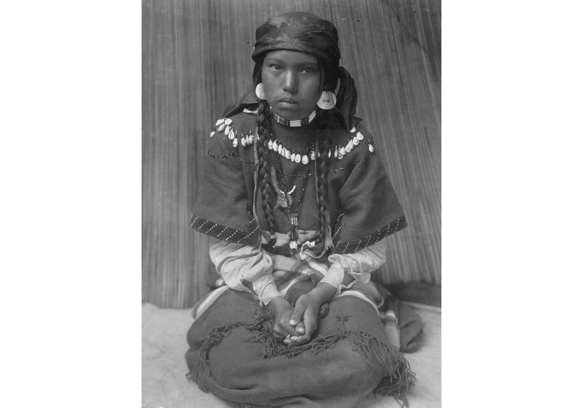 Touch her dress--Kalispel. Kalispel girl sitting on her knees, hands folded in lap, 1910.